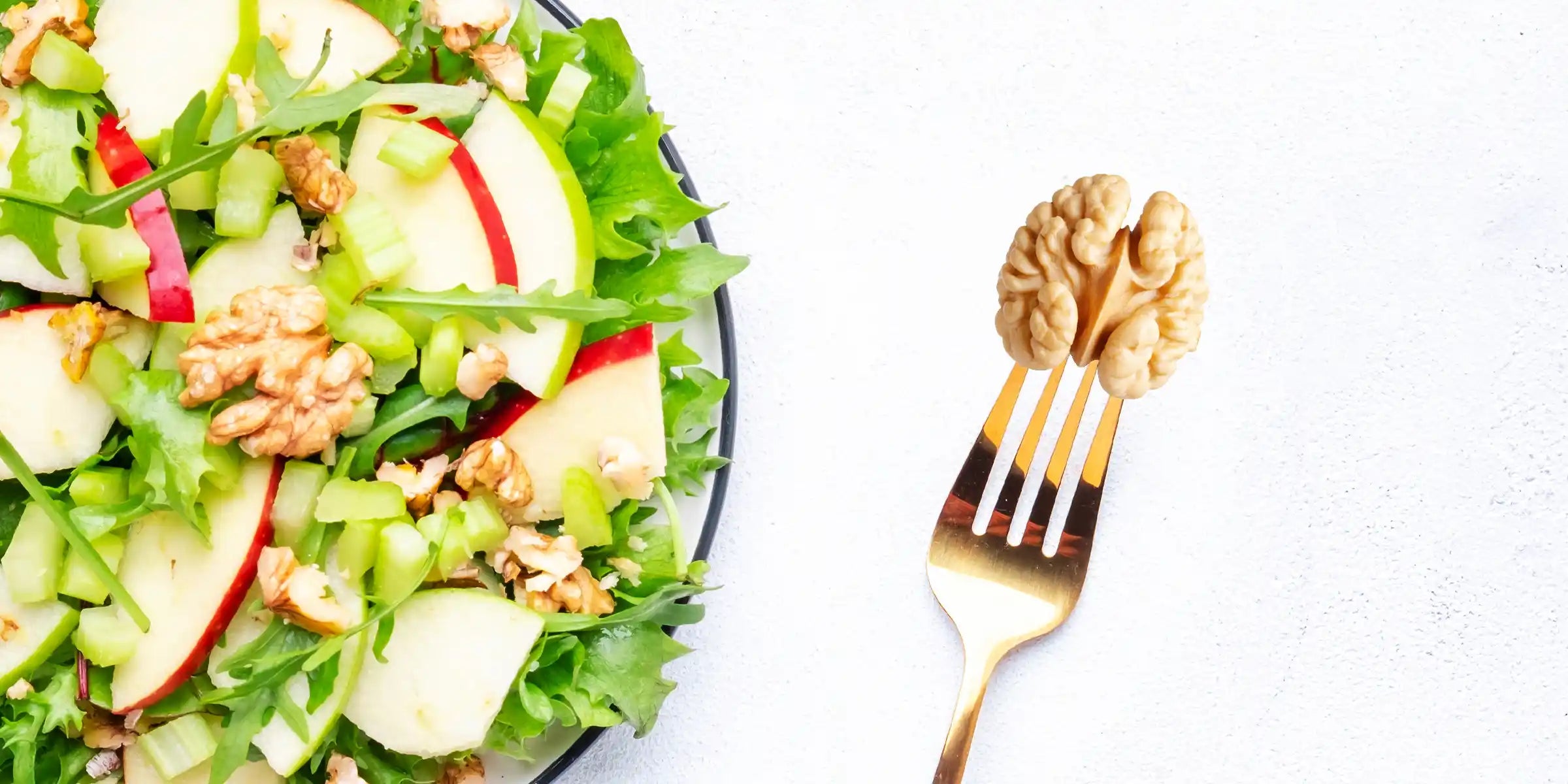 Mixed greens salad with apple slices, celery, and walnuts beside a fork holding a walnut on a light background.
